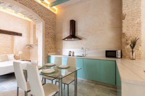 Moderna cocina-comedor con muebles turquesas, mesa de cristal y paredes de ladrillo en apartamento Triana, Sevilla
