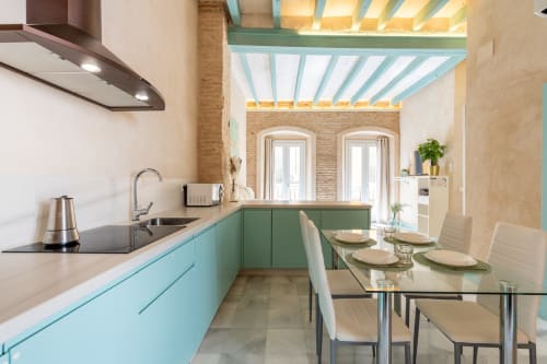Cocina y comedor modernos en apartamento de Sevilla con muebles color turquesa, paredes de ladrillo y mesa de cristal para cuatro personas