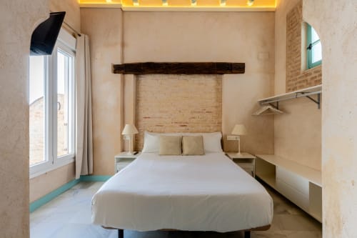 Dormitorio moderno en apartamento de Sevilla con cama blanca, pared de ladrillo visto, viga de madera oscura y vista a la ciudad