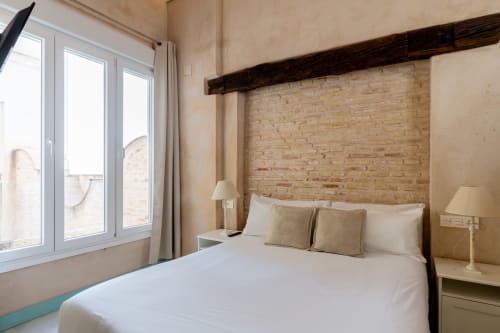 Dormitorio luminoso en apartamento de Sevilla con pared de ladrillo visto, ropa de cama blanca, viga de madera oscura y vistas a la ciudad