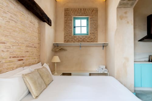 Dormitorio luminoso en apartamento de Sevilla con ladrillo visto, ventana turquesa, ropa de cama blanca y cocina integrada