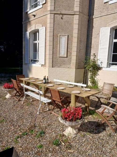 Espace de restauration en plein air à la villa Le château de Mons Boubert avec longue table en bois, couverts et fleurs rouges
