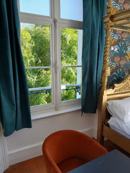 Chambre élégante au château de Mons Boubert avec rideaux bleu-vert, portes-fenêtres, lit à cadre doré ouvragé et vue sur jardin luxuriant