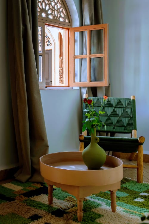 Élégante chambre d'hôtel Marrakech : fenêtre encadrée bois, fauteuil vert, vase céramique fleuri et tapis motifs à Dabachi