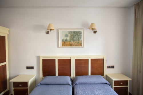 Dos camas individuales con ropa de cama azul en habitación luminosa del Hostal Atalaya, Minas de Ríotinto, con cabeceros de madera y lámparas de pared
