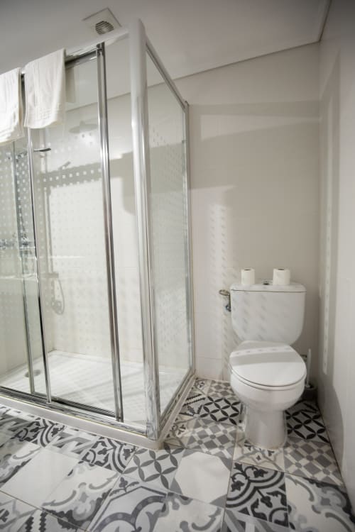 Baño moderno del hotel San Poul en Consuegra con ducha de cristal, inodoro blanco y piso con azulejos estampados