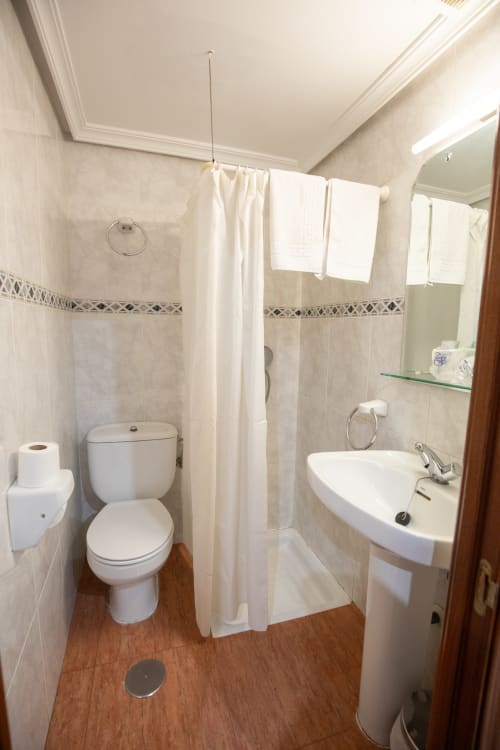 Baño moderno del hotel San Poul en Consuegra con inodoro blanco, lavabo, ducha con cortina y piso de madera