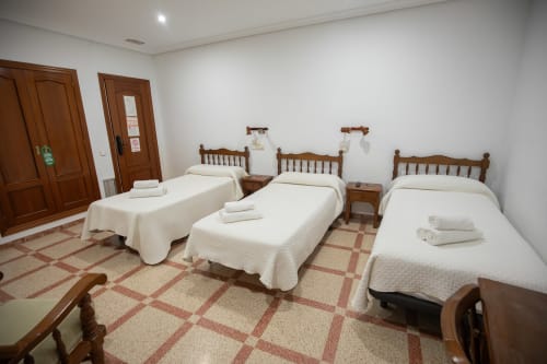 Habitación triple del hotel San Poul en Consuegra con tres camas de madera, ropa de cama blanca y piso de baldosas de terracota.