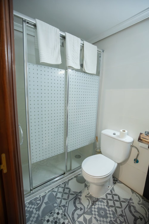 Baño moderno del hotel San Poul en Consuegra con ducha de cristal, toallas blancas y baldosas con patrón