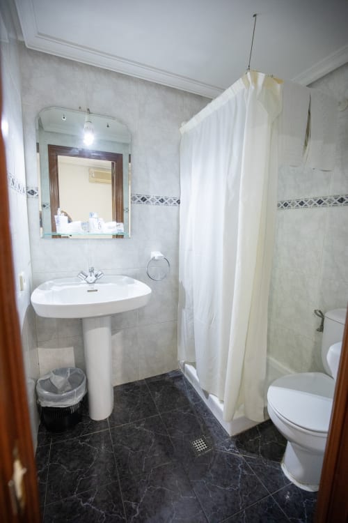 Baño moderno del Hotel San Poul en Consuegra con lavabo de pedestal, inodoro, ducha y piso de baldosas oscuras