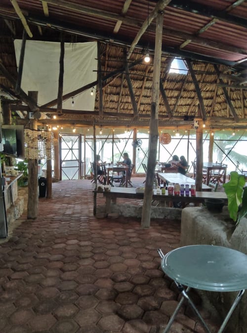 Área común del hostal Cayuco Maya en Bacalar con techo de palapa, vigas de madera y mesas de comedor