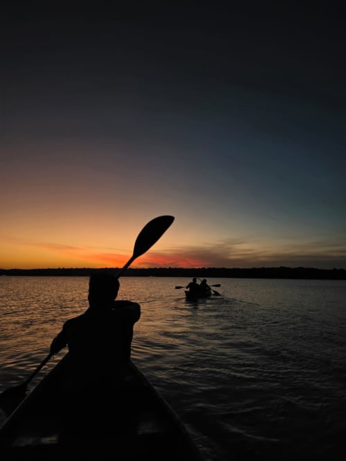 Kayakistas remando en Lago Bacalar al atardecer, silueteados contra el cielo dorado cerca del hostel Cayuco Maya