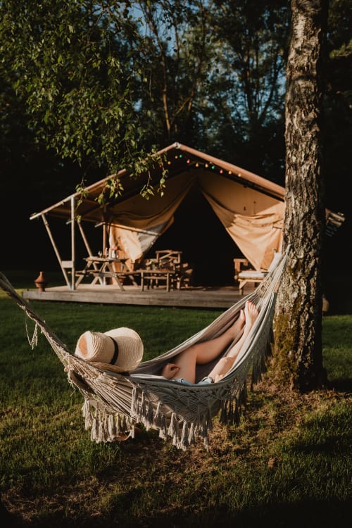 Invité se détendant dans un hamac aux gîtes de Cormenin, avec tente glamping et espace de rassemblement éclairé au feu en arrière-plan
