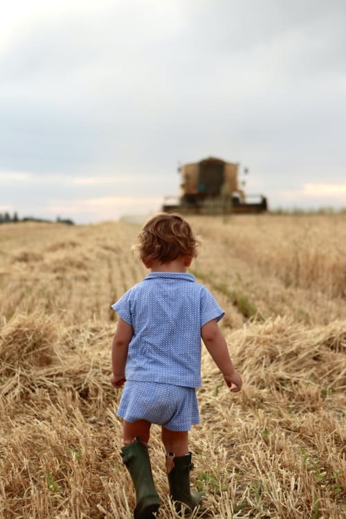 Jeune enfant en robe et bottes bleues courant dans un champ de blé avec une moissonneuse-batteuse en arrière-plan