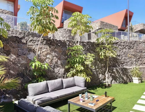 Modern villa patio with stone walls, contemporary red-roofed building, comfortable seating area, lush greenery, and wooden