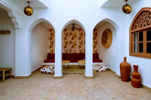 Le salon marocain traditionnel du Riad Dabachi Tala'lou avec alcôves voûtées, sièges rouges, papier peint géométrique et suspension en laiton