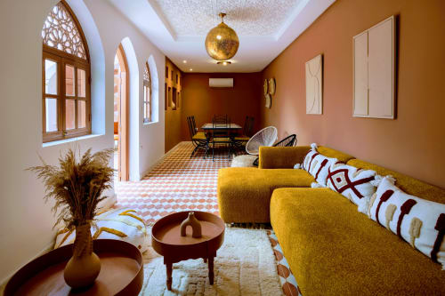 Salon Tala'lou du Riad Dabachi à Marrakech avec canapé moutarde, motifs géométriques, suspension en laiton et fenêtres cintrées.