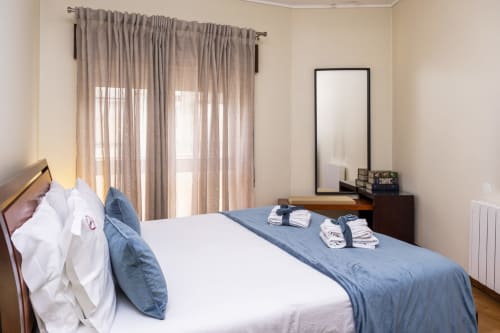 Quarto luminoso nos RH Charming Apartments em Barcelos com roupa de cama branca, almofadas azuis decorativas e cortinas translúcidas com vista
