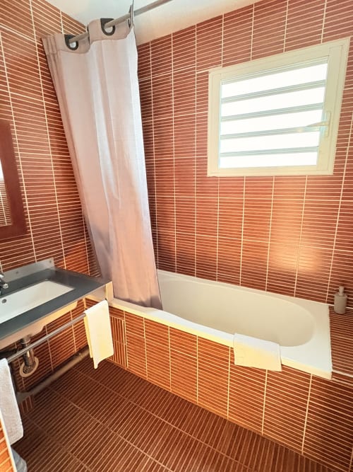 Salle de bain moderne avec carrelage brique rouille, baignoire blanche, rideau de douche rose et fenêtre à l'Hôtel le soleil couchant, St