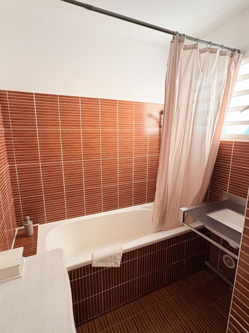 Salle de bain moderne à l'Hôtel le soleil couchant : murs en brique rouge, baignoire blanche, rideau de douche rose et détails sombres