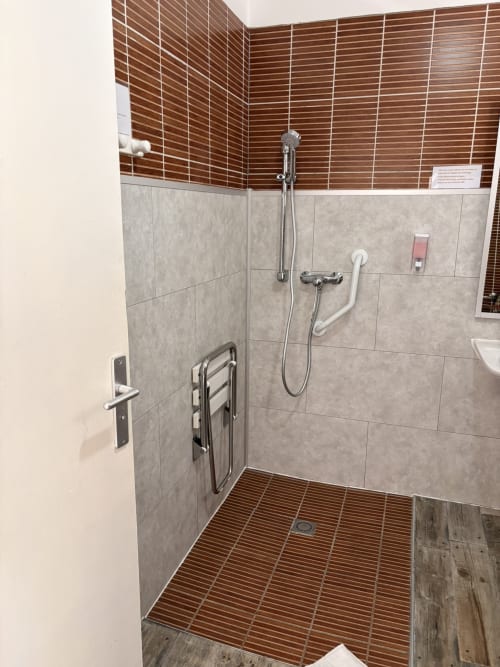 Salle de bains accessible à l'Hôtel le soleil couchant à St Pierre, RE avec barres d'appui, pommeau de douche cascade et sol en bois
