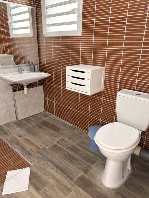 Salle de bains moderne avec carrelage marron, toilettes blanches, lavabo, meuble de rangement et revêtement aspect bois à l'Hôtel le soleil