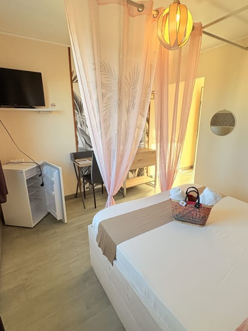 Chambre double de l'Hôtel le Soleil Couchant avec lit, rideaux roses, TV et mini-bar à Saint-Pierre