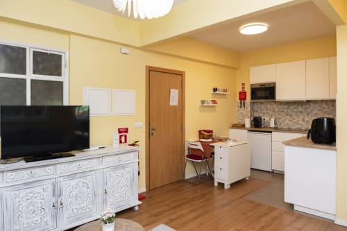Estúdio moderno com paredes amarelas, cozinha equipada, armários brancos, piso de madeira e área de TV no RH Charming