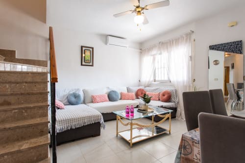 Luminoso salón con sofá en L negro, cojines coloridos, mesa de centro de cristal y ventilador de techo blanco en Casa Bibaziz