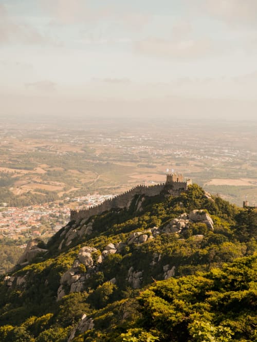 Ruínas de castelo medieval em encosta rochosa acima do vale de Sintra com encostas florestadas e vilarejo abaixo