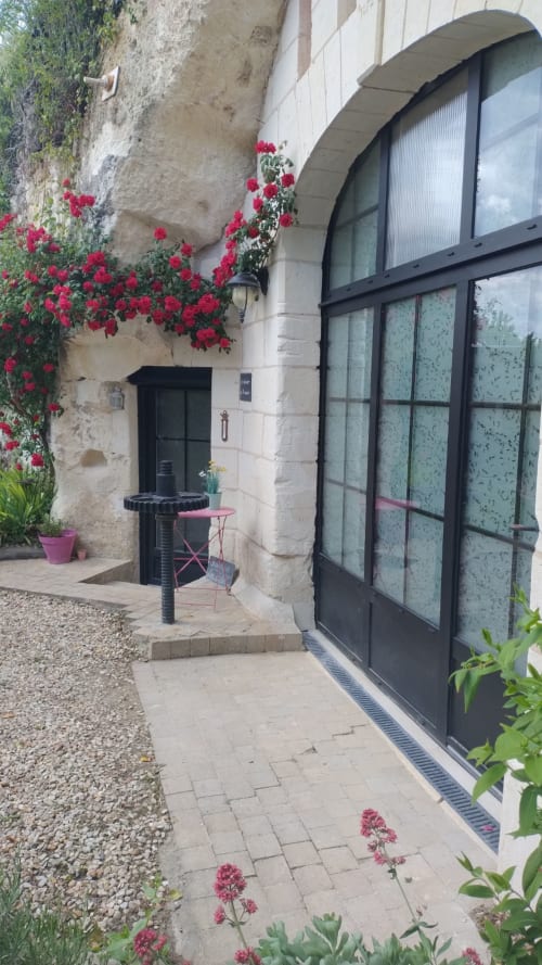 Entrée des Troglos de Beaulieu avec fleurs grimpantes rouges éclatantes, porte noire, fenêtre en arc et terrasse patio avec vue sur le jardin