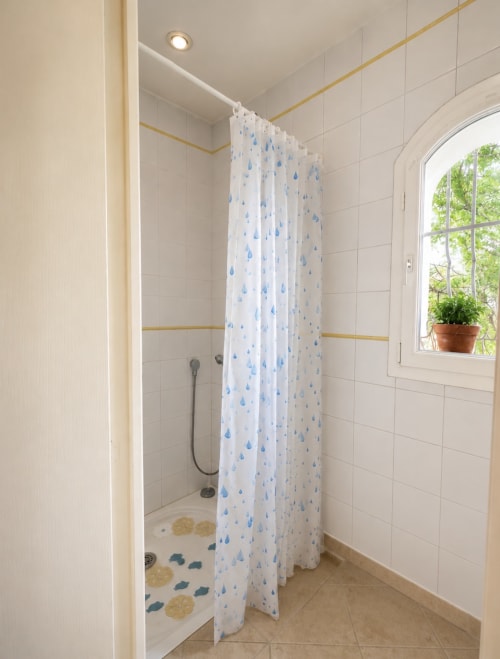 Salle de bains moderne avec carrelage blanc, rideau de douche bleu gouttes de pluie et plante en pot à la Conciergerie Maurin à Arles
