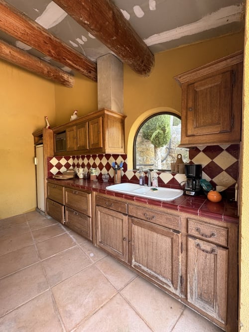 Cuisine rustique aux armoires en bois, murs jaunes, fenêtre en arche et dosseret losanges rouges à la Conciergerie Maurin