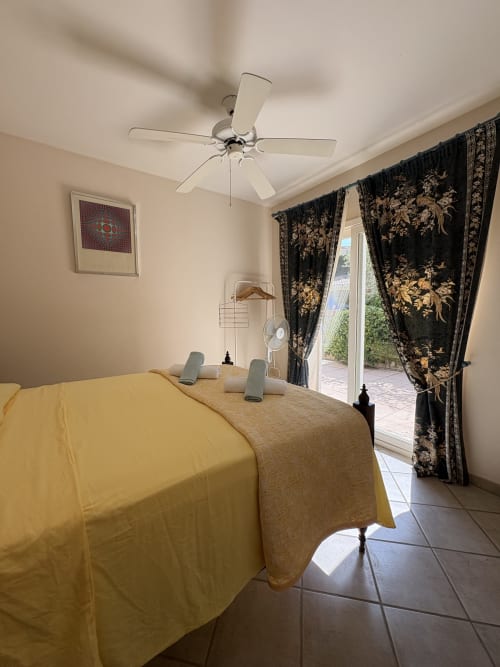 Chambre lumineuse aux draps jaunes, ventilateur de plafond, rideaux à motifs foncés et vue sur jardin à la Conciergerie Maurin à Arles