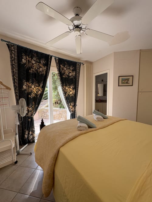 Chambre lumineuse avec literie jaune, rideaux à motifs floraux noirs, ventilateur de plafond et accès à la terrasse à la Conciergerie Maurin à Arles