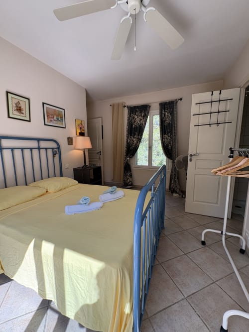 Chambre lumineuse avec lit métallique bleu, linge de lit jaune, ventilateur de plafond blanc, rideaux floraux foncés et carrelage à la Conciergerie