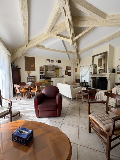 Salon spacieux à la Conciergerie Maurin à Arles avec poutres apparentes, fauteuil bordeaux, cheminée et décor classique