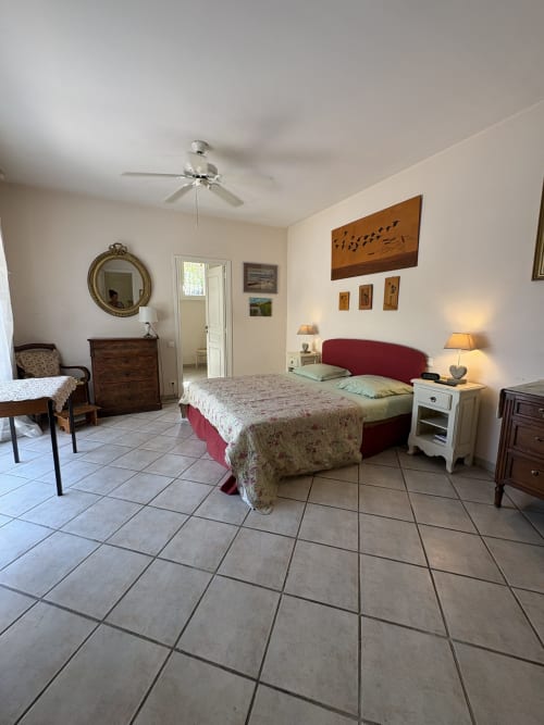 Chambre spacieuse à la Conciergerie Maurin à Arles avec lit rouge, carrelage, mobilier en bois et ventilateur de plafond