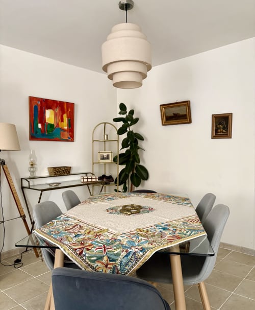 Salle à manger de l'appartement à Arles : nappe à motifs, chaises grises, luminaire suspendu et tableaux encadrés sur murs blancs