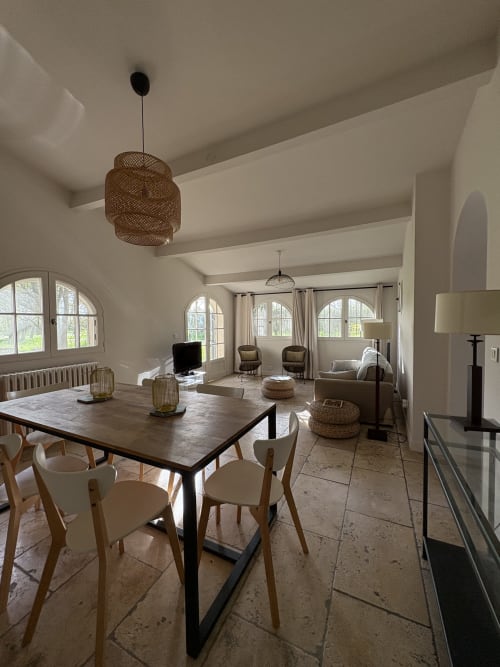 Salle à manger moderne avec table en bois, chaises crème, luminaire suspendu tressé, baies vitrées cintrées donnant sur le jardin du Gîte Château de