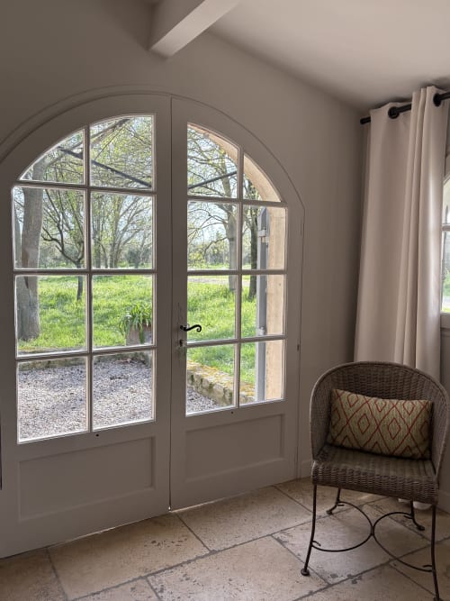 Portes vitrées cintrées ouvrant sur le jardin au Gîte château de Pannis, avec fauteuil en rotin et rideaux crème