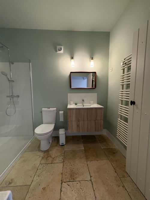Salle de bains moderne au Gîte château de Pannis à Arles avec murs vert sauge, meuble vasque en bois, miroir et carrelage travertin