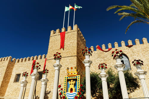 Hostal La Moyareja en Rota, España - fachada de castillo de piedra con torres almenadas, banderas decorativas y cinta roja festiva