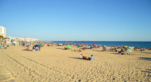 Playa concurrida de Rota con sombrillas coloridas, bañistas y edificios costeros bajo cielo azul despejado