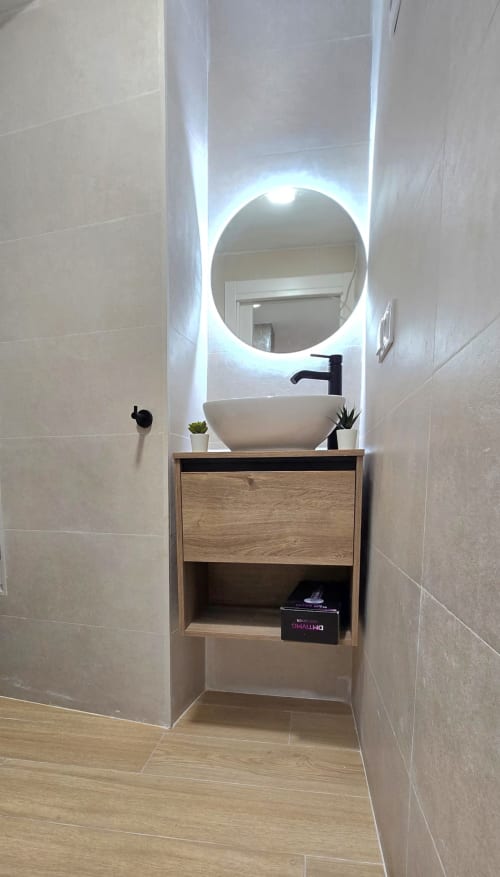 Lavabo moderno con lavamanos de cerámica blanca, grifo negro y espejo redondo iluminado en apartamento de Murcia