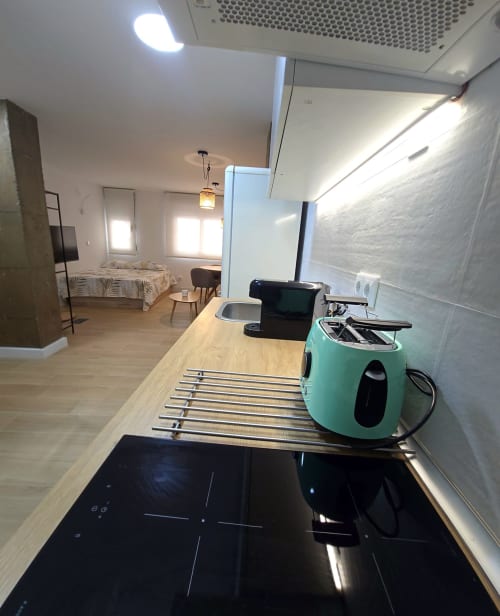 Moderno kitchenette de estudio en Murcia con placa de inducción, hervidor verde menta y dormitorio abierto con luz natural