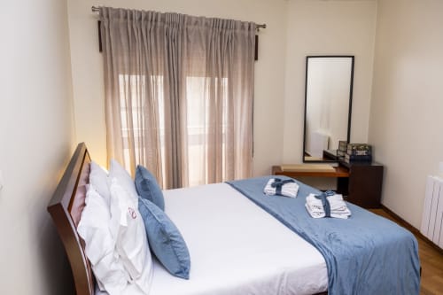 Quarto luminoso nos RH Charming Apartments em Braga com detalhes azuis, cama de madeira, espelho e cortinas translúcidas