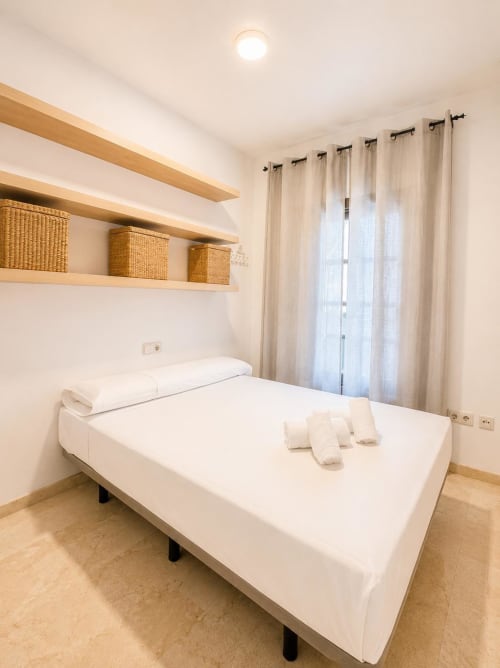 Dormitorio luminoso en apartamento de Málaga con ropa de cama blanca, estantes de madera, cortinas beige e iluminación cálida