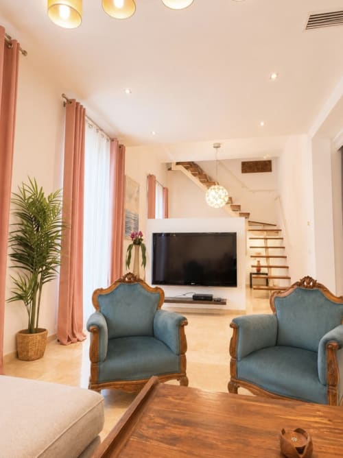Moderno salón dúplex en Málaga con sillones azules, TV de pantalla plana, escalera de madera y cortinas color coral