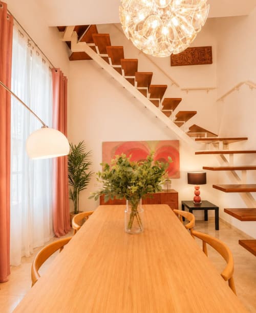 Comedor moderno en dúplex de Málaga con mesa de madera, escalera flotante, iluminación colgante y obra de arte contemporáneo.